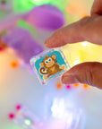 Main tenant un cube lumineux Zany Zoo de Glo Pals illustré d’un singe sur fond turquoise, au-dessus d’un bac de jeu sensoriel.