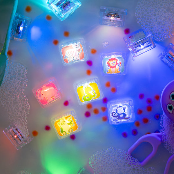 Cubes lumineux Zany Zoo de Glo Pals allumés dans un bac d’eau avec mousse, petits pompons colorés et outils de jeu violets.