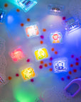 Cubes lumineux Zany Zoo de Glo Pals allumés dans un bac d’eau avec mousse, petits pompons colorés et outils de jeu violets.