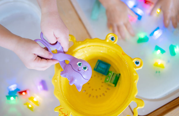Mains manipulant un outil violet au-dessus d’une passoire jaune en forme de crabe, dans un bac d’eau avec cubes lumineux Glo Pals.
